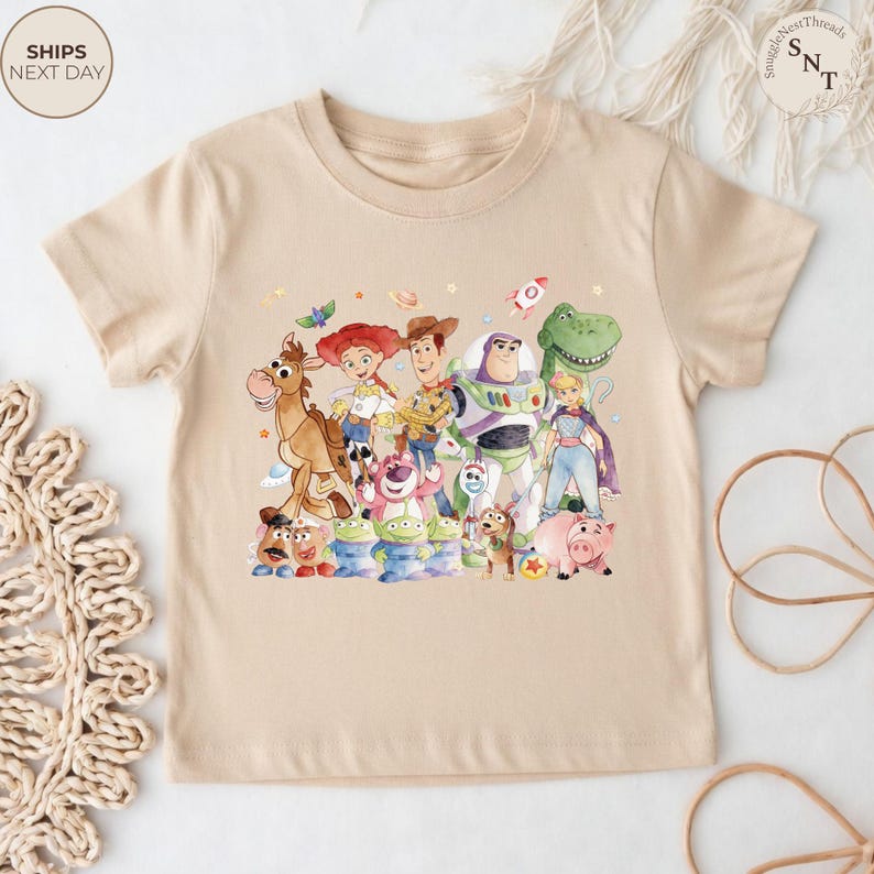 May include: A beige t-shirt featuring a colorful watercolor illustration of characters from the Toy Story movie franchise. The design includes Woody, Buzz Lightyear, Jessie, and other characters. The shirt has short sleeves and a crew neck.