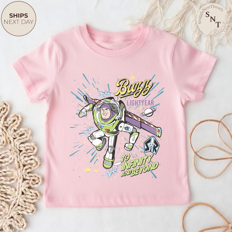 Puede incluir: Una camiseta rosa claro con un dise&ntilde;o de Buzz Lightyear. El dise&ntilde;o incluye el texto "Buzz Lightyear" y "To Infinity and Beyond". La imagen es de estilo caricatura con colores vibrantes.