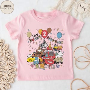 Puede incluir: Una camiseta rosa claro para ni&ntilde;os peque&ntilde;os con un gr&aacute;fico colorido de coches de dibujos animados, globos y decoraciones de cumplea&ntilde;os. El dise&ntilde;o incluye el nombre "LILY" y el n&uacute;mero "2", con las palabras "HAPPY BIRTHDAY".