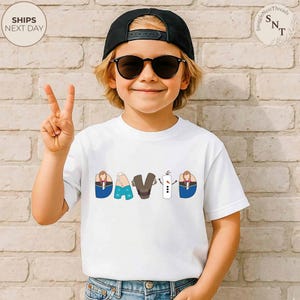K&ouml;nnte beinhalten: Ein wei&szlig;es T-Shirt mit dem Namen "DAVID" in einem farbenfrohen Cartoon-Stil aufgedruckt. Die Buchstaben sind mit Bildern von Charakteren gestaltet. Das Kind tr&auml;gt eine schwarze Kappe und eine Sonnenbrille.
