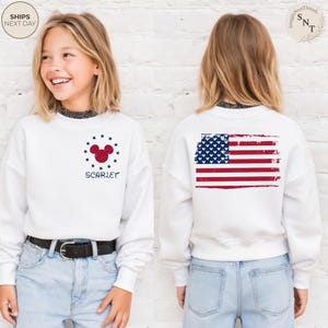 May include: White sweatshirt featuring a red Mickey Mouse head surrounded by blue stars and the name "Scarlet". The back of the sweatshirt displays a distressed American flag design. The sweatshirt is worn with blue jeans.