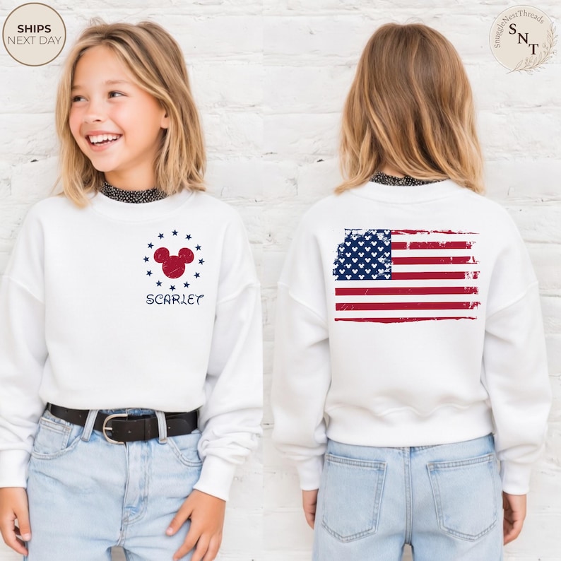May include: White sweatshirt featuring a red Mickey Mouse head surrounded by blue stars and the name "Scarlet". The back of the sweatshirt displays a distressed American flag design. The sweatshirt is worn with blue jeans.