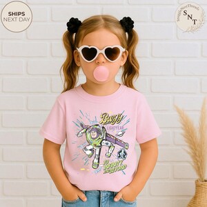 Puede incluir: Camiseta rosa claro con un dise&ntilde;o de Buzz Lightyear y las frases "Buzz Lightyear" y "To Infinity and Beyond". La camiseta la lleva un ni&ntilde;o con gafas de sol en forma de coraz&oacute;n.