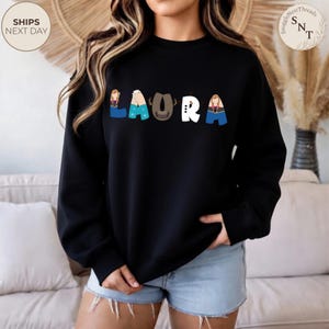 K&ouml;nnte beinhalten: Schwarzes Sweatshirt mit Rundhalsausschnitt und dem Namen "LAURA" in bunten Buchstaben im Cartoon-Stil. Die Buchstaben sind mit Bildern von Charakteren gestaltet. Das Sweatshirt hat eine lockere Passform und wird mit Jeansshorts getragen.