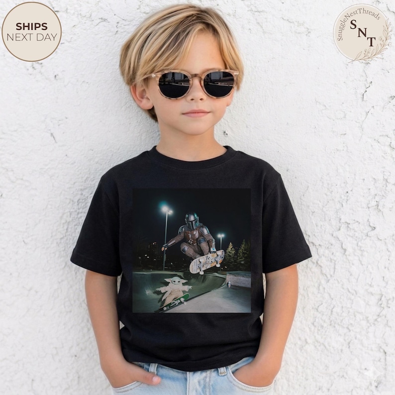 May include: A black t-shirt featuring a graphic of a character from The Mandalorian skateboarding, with Grogu on a skateboard below. The shirt is worn by a child wearing sunglasses. The text "SHIPS NEXT DAY" is in a circle on the upper left.
