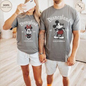 Pu&ograve; includere: T-shirt grigie con grafiche di Disneyland con Topolino e Minnie. Le magliette mostrano "Disneyland" sopra le illustrazioni dei personaggi, con "est. 1955" e "CALIFORNIA" sotto. La coppia indossa pantaloncini bianchi.