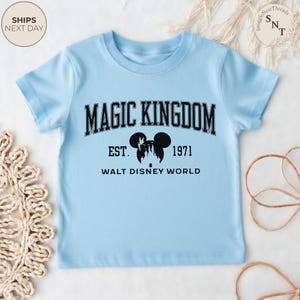 May include: A light blue t-shirt featuring the text "MAGIC KINGDOM" in black, with "EST. 1971" and "WALT DISNEY WORLD" below. A black silhouette of Mickey Mouse ears and a castle is in the center. The shirt is short-sleeved.