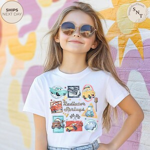 May include: A white t-shirt featuring colorful cartoon car designs and the text "Radiator Springs". The shirt has a crew neck and short sleeves. The design includes various car characters in stamp-like frames.