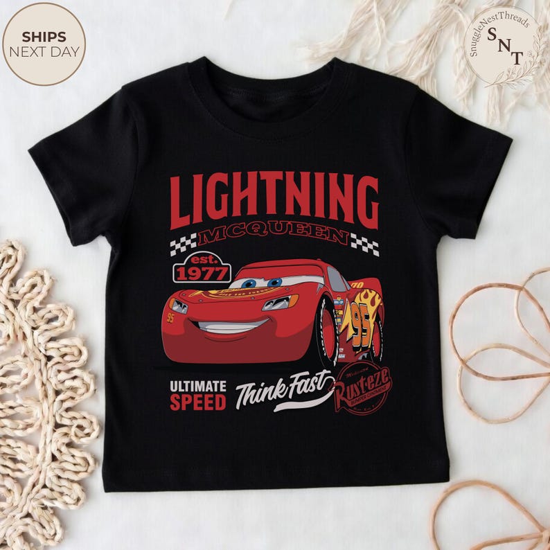 Op de afbeelding: Zwart T-shirt met een Lightning McQueen-ontwerp uit de film Cars. Het ontwerp bevat de tekst "LIGHTNING MCQUEEN" in het rood, het jaar "est. 1977" en de zinnen "ULTIMATE SPEED" en "Think Fast". Er is ook een cartoonafbeelding van de rode raceauto.