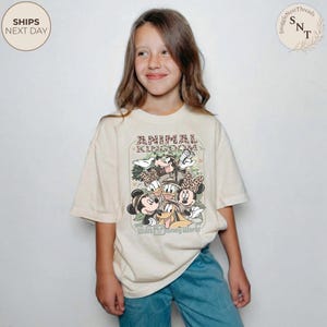Pu&ograve; includere: T-shirt color crema con una grafica di Topolino, Minnie, Paperino, Paperina e Pippo. La maglietta riporta le scritte "ANIMAL KINGDOM" e "Walt Disney World". La maglietta &egrave; indossata da una persona.