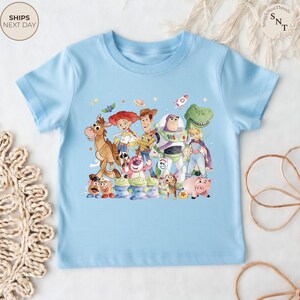 May include: A light blue t-shirt featuring a colorful graphic of characters from the animated movie Toy Story. The design includes Woody, Buzz Lightyear, Jessie, and other characters. The shirt has short sleeves and a crew neck.