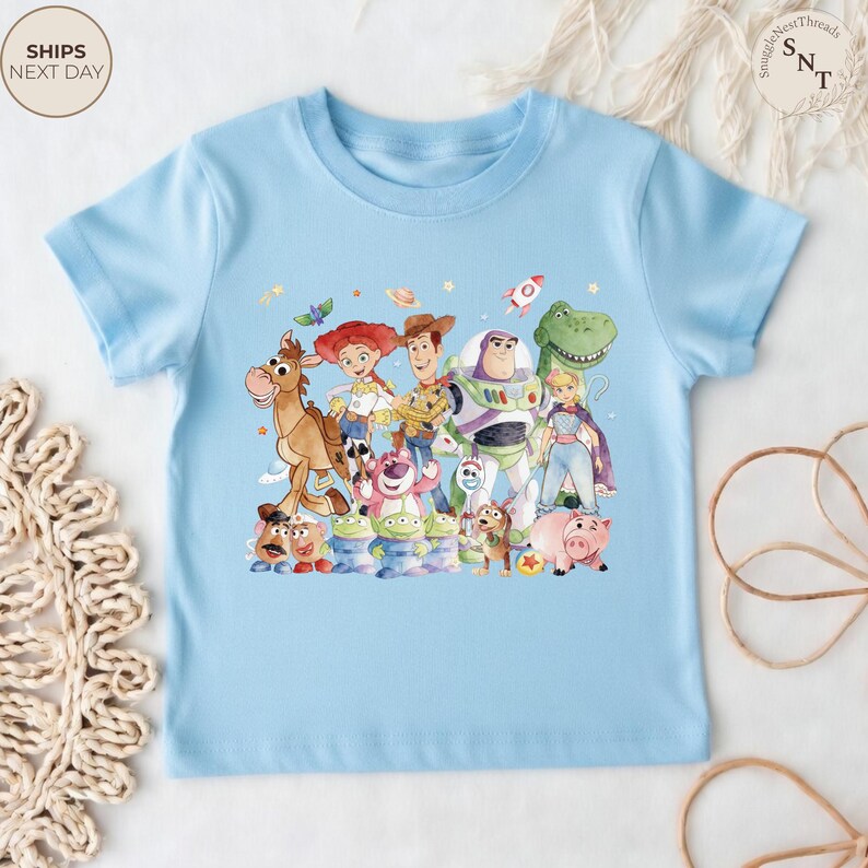 May include: A light blue t-shirt featuring a colorful graphic of characters from the animated movie Toy Story. The design includes Woody, Buzz Lightyear, Jessie, and other characters. The shirt has short sleeves and a crew neck.