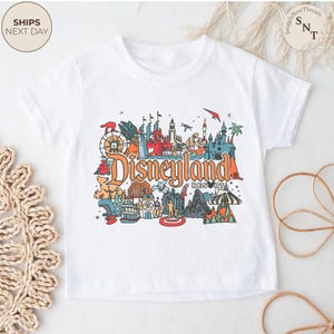 May include: White t-shirt featuring a colorful Disneyland Resort graphic. The design includes iconic landmarks and the Disneyland Resort logo in orange. The shirt is a short-sleeved crew neck.
