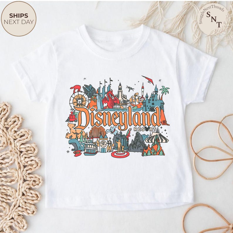 May include: White t-shirt featuring a colorful Disneyland Resort graphic. The design includes iconic landmarks and the Disneyland Resort logo in orange. The shirt is a short-sleeved crew neck.