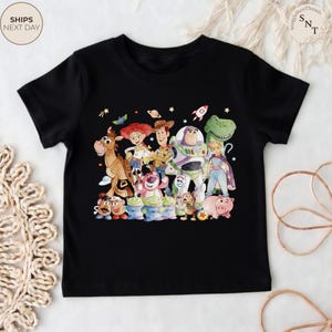 May include: Black t-shirt featuring a colorful graphic of characters from the animated film "Toy Story." The design includes Woody, Buzz Lightyear, Jessie, and other beloved characters. The shirt is a crew neck style.