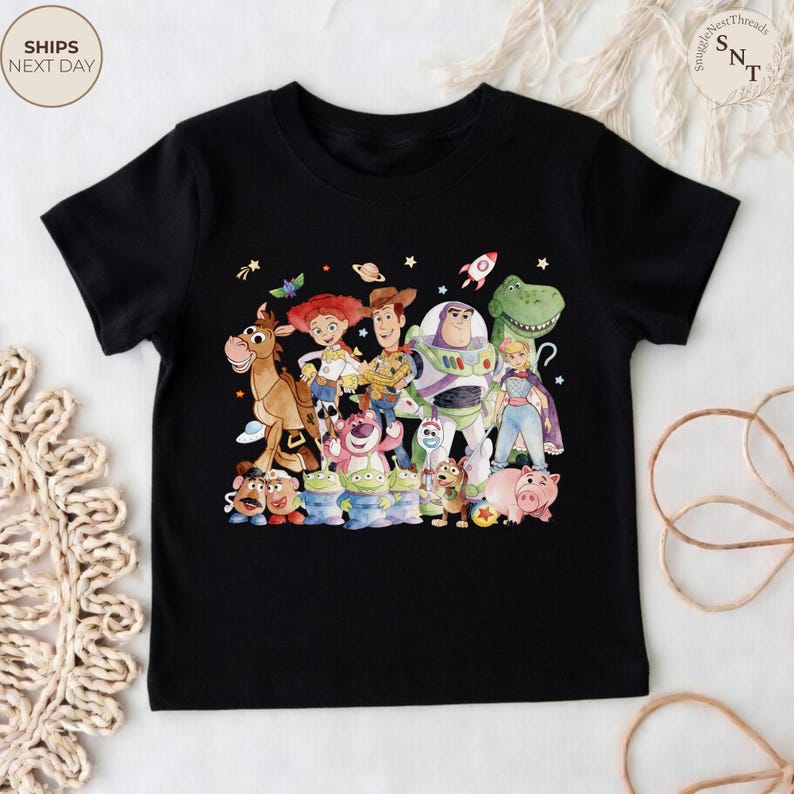May include: Black t-shirt featuring a colorful graphic of characters from the animated film "Toy Story." The design includes Woody, Buzz Lightyear, Jessie, and other beloved characters. The shirt is a crew neck style.