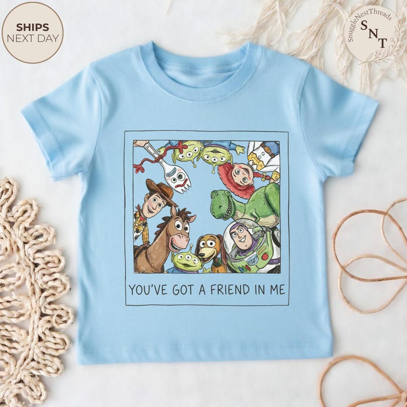 May include: Light blue short-sleeved t-shirt featuring a colorful graphic of characters from the movie "Toy Story". The graphic is framed in a black rectangle and includes the text "YOU'VE GOT A FRIEND IN ME".