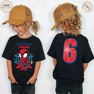 Puede incluir: Camiseta negra con un dise&ntilde;o de Spider-Man y el texto "THE AMAZING JACOB" en la parte delantera. La parte trasera de la camiseta muestra un gran n&uacute;mero "6" con un dise&ntilde;o de telara&ntilde;a. El ni&ntilde;o lleva una gorra de camionero marr&oacute;n.