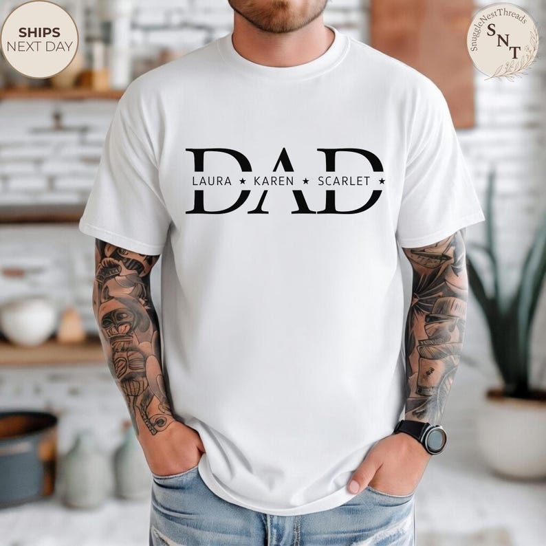 Puede incluir: Camiseta blanca con la palabra "DAD" en letras negras y negritas. Debajo, los nombres "LAURA", "KAREN" y "SCARLET" est&aacute;n impresos con estrellas. La camiseta es de estilo casual.