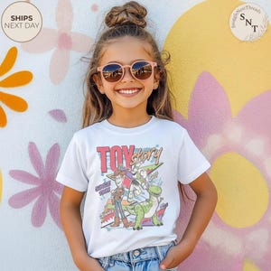 Może przedstawiać: Biała koszulka z logo Toy Story i postaciami Woody'ego, Buzza Astrala i Rexa. Koszulka ma napis "Toy Story" i "Since 1995" w kolorowym, retro designie. Dziecko ma na sobie r&oacute;żowe okulary przeciwsłoneczne.