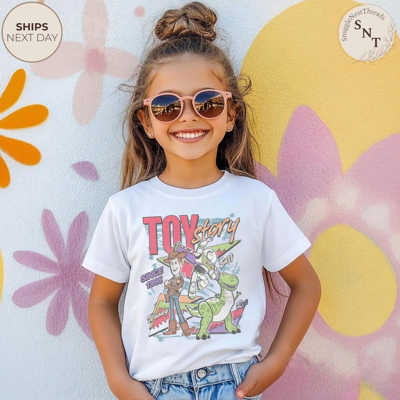 Może przedstawiać: Biała koszulka z logo Toy Story i postaciami Woody'ego, Buzza Astrala i Rexa. Koszulka ma napis "Toy Story" i "Since 1995" w kolorowym, retro designie. Dziecko ma na sobie r&oacute;żowe okulary przeciwsłoneczne.