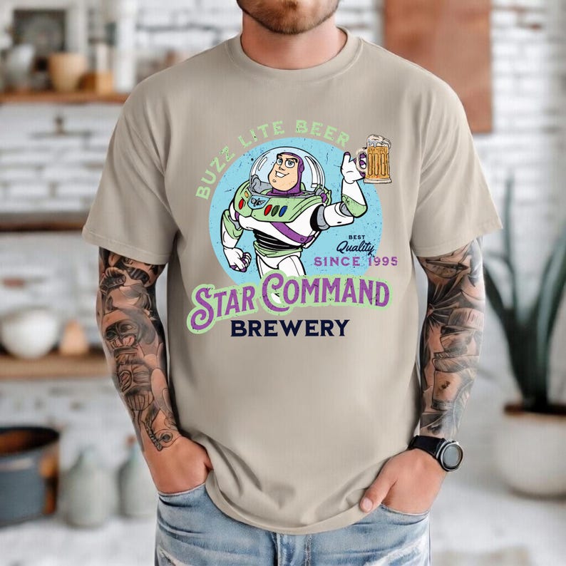 Puede incluir: Camiseta beige con un dise&ntilde;o de dibujos animados de Buzz Lightyear sosteniendo una jarra de cerveza. El dise&ntilde;o incluye el texto "Buzz Lite Beer" y "Star Command Brewery" con "Best Quality Since 1995". La camiseta tiene cuello redondo y mangas cortas.