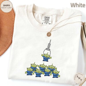 May include: White Comfort Colors t-shirt featuring a graphic of the Little Green Men from Toy Story being lifted by a claw machine. The aliens are wearing blue uniforms. A gold necklace is draped across the shirt.