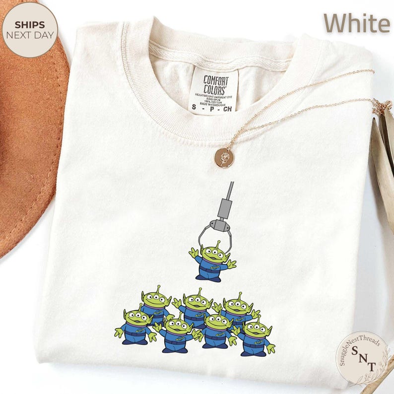 May include: White Comfort Colors t-shirt featuring a graphic of the Little Green Men from Toy Story being lifted by a claw machine. The aliens are wearing blue uniforms. A gold necklace is draped across the shirt.