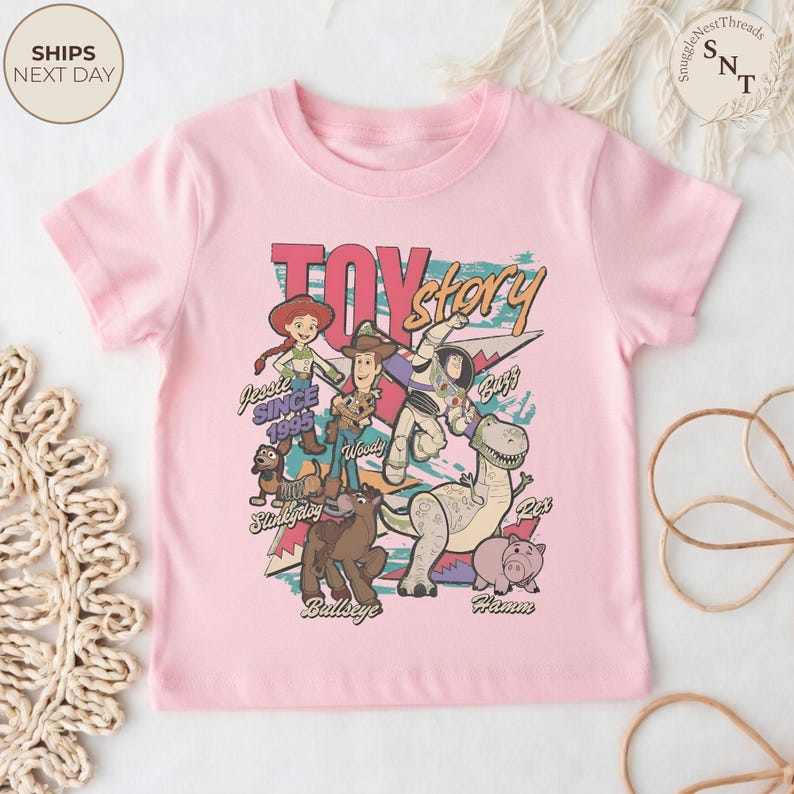 Puede incluir: Una camiseta de manga corta de color rosa claro con un estampado gr&aacute;fico de los personajes de Toy Story, incluyendo a Jessie, Woody, Buzz Lightyear, Slinky Dog, Bullseye, Rex y Hamm. El texto dice Toy Story, Jessie Since 1995, Slinky Dog, Woody, Buzz, Bullseye y Hamm.