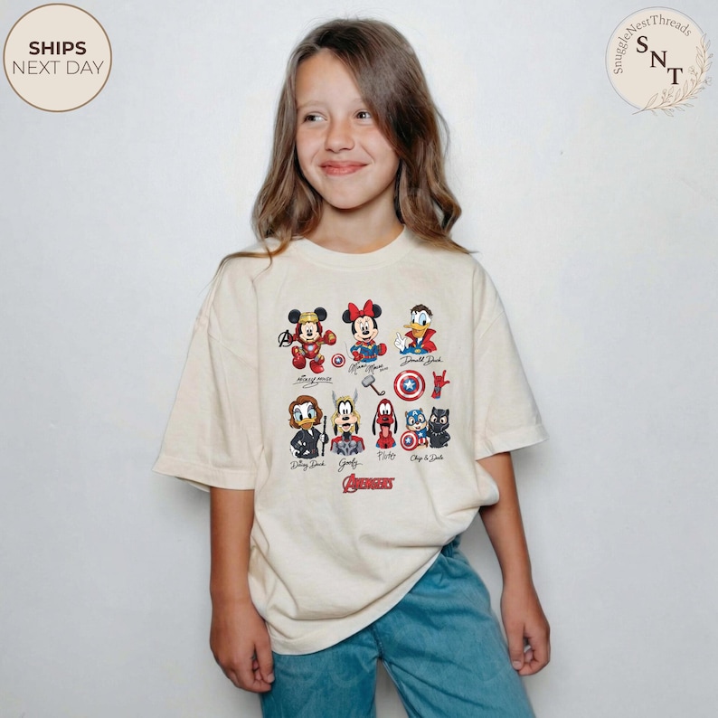 May include: A cream-colored t-shirt featuring cartoon characters, including Mickey Mouse, Minnie Mouse, and Donald Duck, with the word "Avengers" in red. The shirt is worn by a person.
