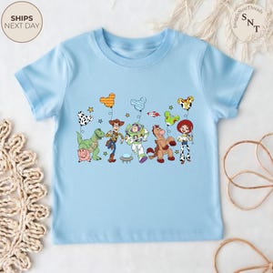 May include: A light blue t-shirt featuring a colorful graphic of Toy Story characters, including Woody, Buzz Lightyear, and Jessie. The design includes balloons and stars. The shirt has short sleeves and a round neckline.