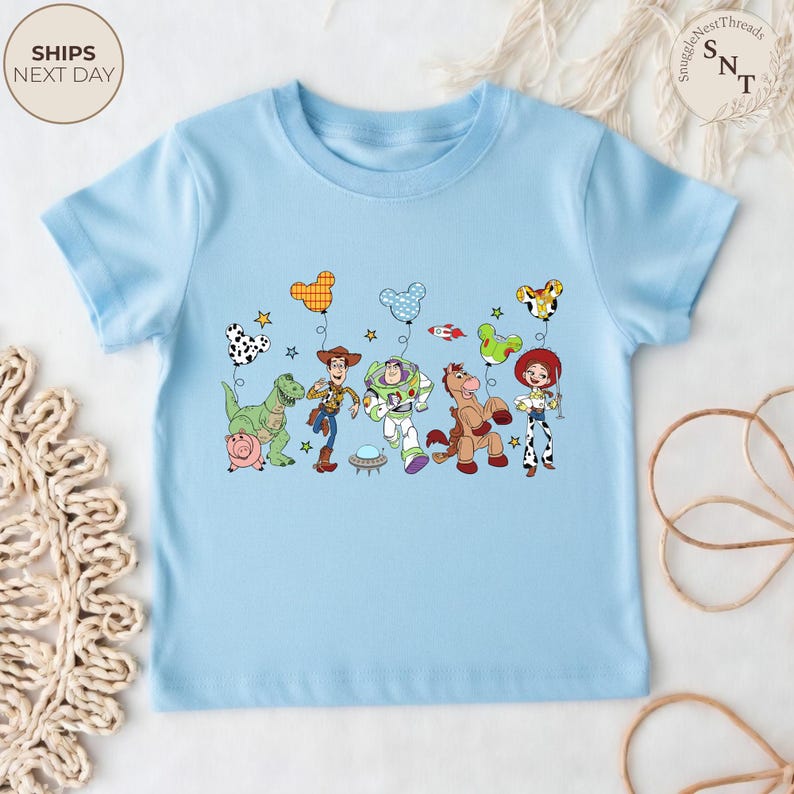 May include: A light blue t-shirt featuring a colorful graphic of Toy Story characters, including Woody, Buzz Lightyear, and Jessie. The design includes balloons and stars. The shirt has short sleeves and a round neckline.
