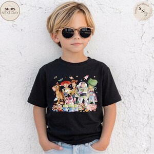 May include: A black t-shirt featuring a colorful graphic of characters from the animated movie "Toy Story." The design includes Woody, Buzz Lightyear, and other beloved characters. The shirt is worn by a child wearing sunglasses.