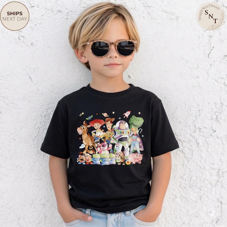 May include: A black t-shirt featuring a colorful graphic of characters from the animated movie "Toy Story." The design includes Woody, Buzz Lightyear, and other beloved characters. The shirt is worn by a child wearing sunglasses.