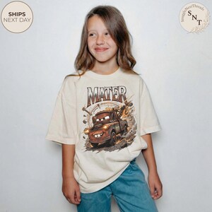 Puede incluir: Camiseta color crema con una imagen de dibujos animados de Mate de la pel&iacute;cula Cars, con el nombre "MATER" encima. El gr&aacute;fico es en tonos marrones, naranjas y amarillos. La camiseta la lleva una persona.