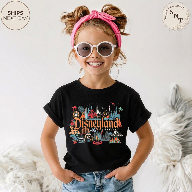 May include: A black t-shirt featuring a colorful Disneyland Resort graphic. The design includes various iconic landmarks and imagery associated with the theme park. The shirt is worn by a child with a pink headband and sunglasses.