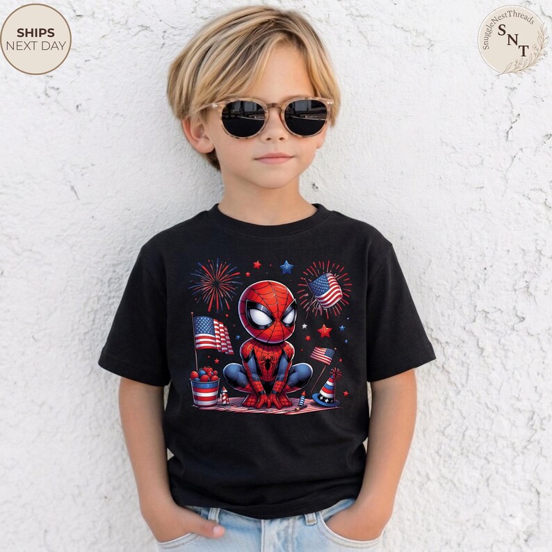 May include: A black short-sleeved t-shirt featuring a graphic of a red and blue superhero character crouching. The design includes American flags, fireworks, a bucket of berries, and a party hat, all rendered in a patriotic red, white, and blue color scheme.