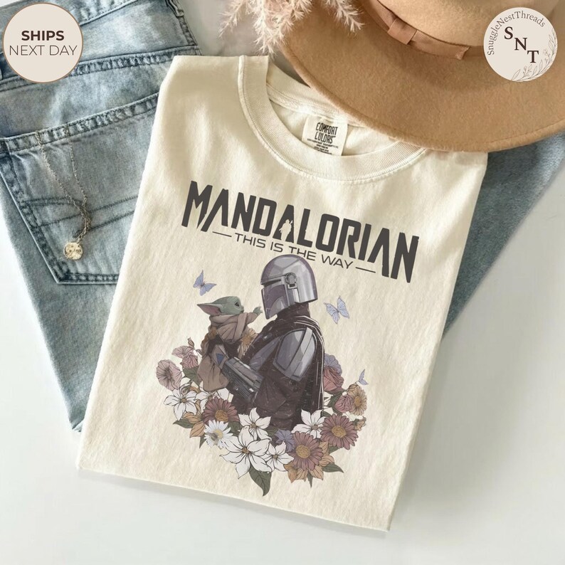 Puede incluir: Una camiseta de color crema con un gr&aacute;fico del Mandaloriano sosteniendo a Grogu, rodeado de varias flores y mariposas. El texto sobre el gr&aacute;fico dice "MANDALORIAN" y "THIS IS THE WAY" en una fuente oscura, negrita y sin remates.