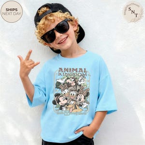Pu&ograve; includere: Una maglietta azzurra con una grafica di personaggi Disney, tra cui Topolino e Minnie, Paperino e Pippo, con la scritta "ANIMAL KINGDOM" e "Walt Disney World". La maglietta &egrave; indossata da un bambino con occhiali da sole e un berretto nero.