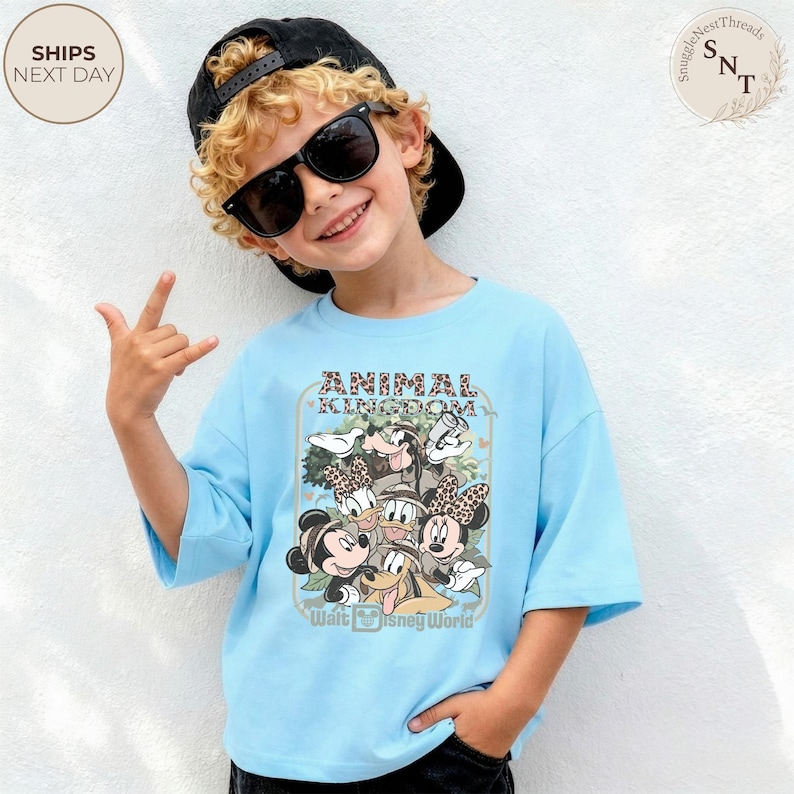 Pu&ograve; includere: Una maglietta azzurra con una grafica di personaggi Disney, tra cui Topolino e Minnie, Paperino e Pippo, con la scritta "ANIMAL KINGDOM" e "Walt Disney World". La maglietta &egrave; indossata da un bambino con occhiali da sole e un berretto nero.