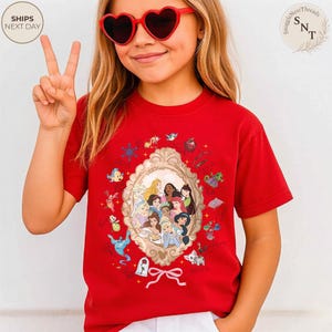 May include: A red t-shirt featuring a framed illustration of Disney princesses. The shirt has a crew neck and short sleeves. The image includes various Disney characters and icons. The model is wearing heart-shaped sunglasses.