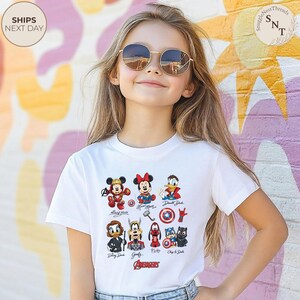 May include: A white t-shirt featuring cartoon characters including Mickey Mouse, Minnie Mouse, and Donald Duck, with the word "AVENGERS" at the bottom. The shirt is worn by a young person wearing sunglasses.