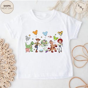 May include: White t-shirt featuring a colorful graphic of Toy Story characters, including Woody, Buzz Lightyear, and Jessie, with balloons. The design includes a dinosaur, a horse, and a pig. The shirt has short sleeves.