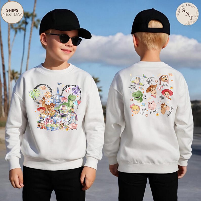 Puede incluir: Una sudadera blanca de manga larga con un gr&aacute;fico colorido de personajes de Toy Story y un castillo en la parte delantera, y una colecci&oacute;n de rostros de personajes con sus nombres impresos en la parte trasera, junto a pantal&oacute;n y gorra negros.