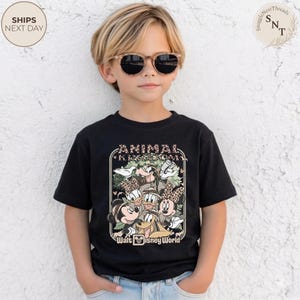 Pu&ograve; includere: T-shirt nero con una grafica colorata dei personaggi Disney, tra cui Topolino e Minnie, Paperino e Paperina, e Pippo. La maglietta presenta la scritta "ANIMAL KINGDOM" e "Walt Disney World".