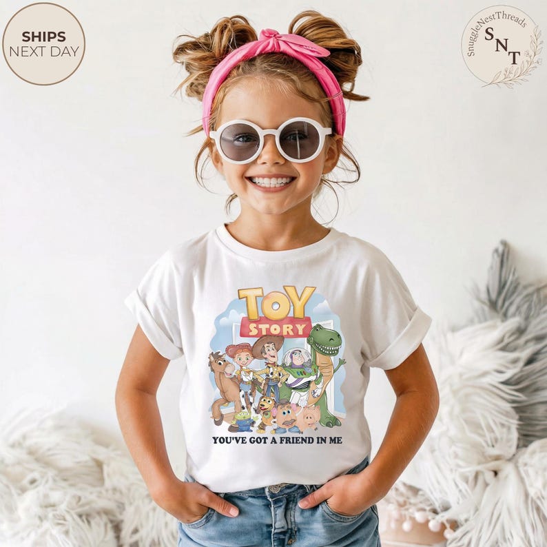 K&ouml;nnte beinhalten: Wei&szlig;es T-Shirt mit einer bunten Grafik von "Toy Story"-Charakteren. Das Shirt zeigt den Text "TOY STORY" und "YOU'VE GOT A FRIEND IN ME". Das Kind tr&auml;gt ein rosa Stirnband und eine Sonnenbrille.