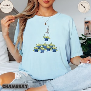May include: Light blue t-shirt featuring a graphic of green aliens from a claw machine. The aliens are wearing blue uniforms. The shirt has the text "CHAMBRAY" at the bottom and "SHIPS NEXT DAY" on the top left.