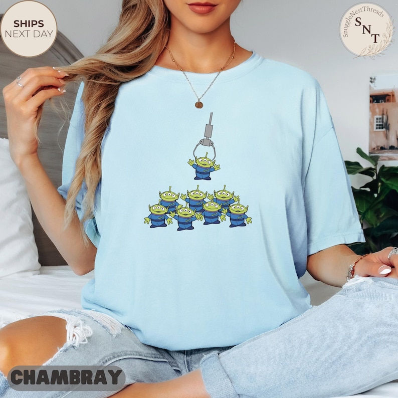 May include: Light blue t-shirt featuring a graphic of green aliens from a claw machine. The aliens are wearing blue uniforms. The shirt has the text "CHAMBRAY" at the bottom and "SHIPS NEXT DAY" on the top left.