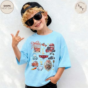May include: Light blue t-shirt featuring a colorful graphic of various Cars movie characters. The design includes images of race cars, trucks, and other vehicles, with the word "Cars" prominently displayed. The shirt is worn by a child.