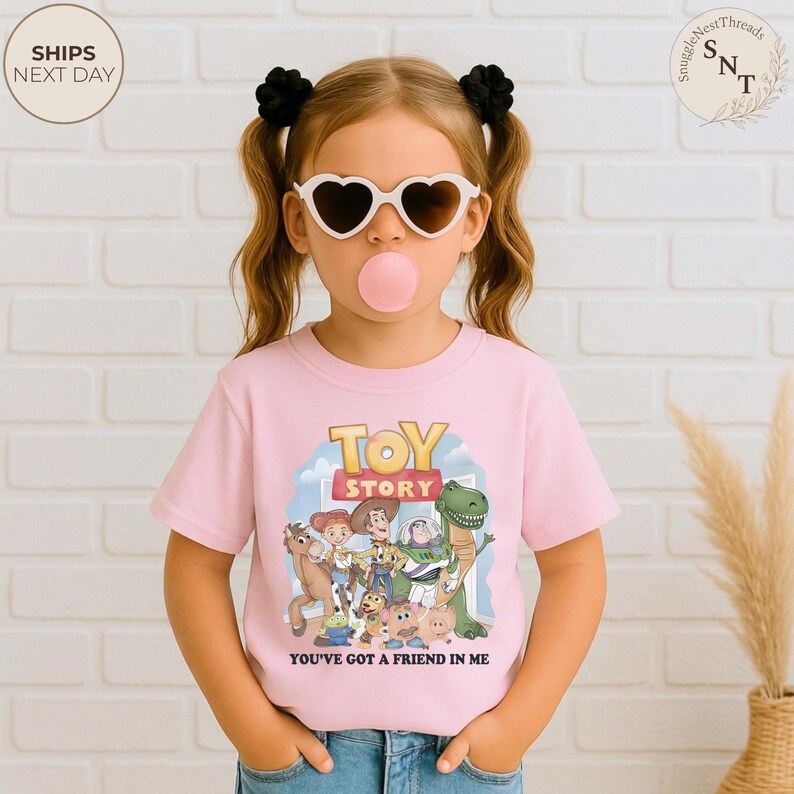K&ouml;nnte beinhalten: Ein rosa T-Shirt mit dem "Toy Story"-Logo und den Figuren sowie dem Schriftzug "YOU'VE GOT A FRIEND IN ME". Das T-Shirt wird von einem Kind mit herzf&ouml;rmiger Sonnenbrille und schwarzen Haargummis getragen.